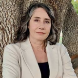 A person with long salt-and-pepper hair, a black shirt, and a beige blazer leans against a tree